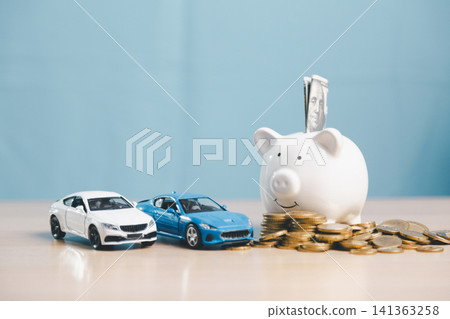 Piggy bank with cash, white and blue toy cars, and coins on desk, representing car savings, auto loan planning, vehicle finance, down payment, and purchase budget. Piggy bank with cash, white and blue toy cars, and coins on desk, representing car savings, auto loan planning, vehicle finance, down payment, and purchase budget. 141363258