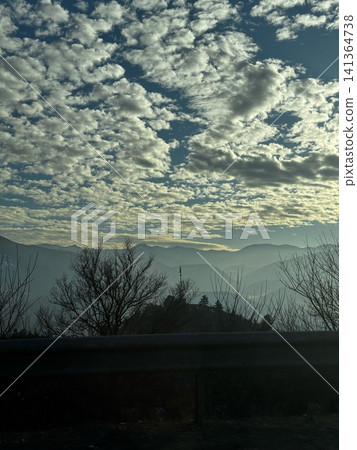 Mountain layers and cloudy sky viewed from roadside with trees in foreground. Mountain layers and cloudy sky viewed from roadside with trees in foreground. 141364738