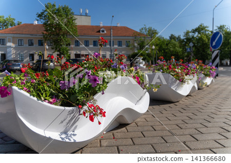 Colorful flowers fill sculptural white urban planters along a sunny city street, with historic architecture, parked cars and a blue traffic sign in the background. Colorful flowers fill sculptural white urban planters along a sunny city street, with historic architecture, parked cars and a blue traffic sign in the background. 141366680