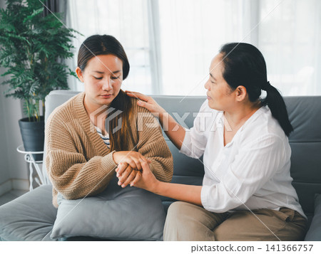 Middle aged Asian woman comforting sad adult daughter by holding hands on sofa at home, concept of emotional support, family care, empathy, counseling, and trust. Middle aged Asian woman comforting sad adult daughter by holding hands on sofa at home, concept of emotional support, family care, empathy, counseling, and trust. 141366757