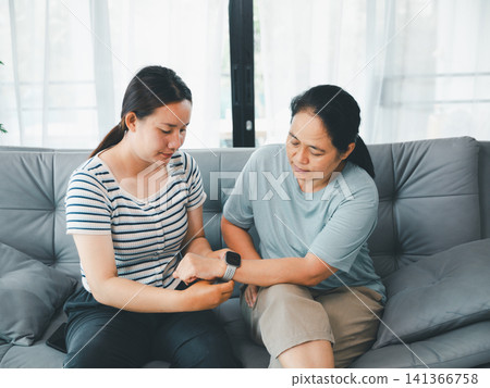 Asian mother and adult daughter checking smartwatch health tracker on sofa at home, concept of wearable technology, family care, digital healthcare, wellness, and support. Asian mother and adult daughter checking smartwatch health tracker on sofa at home, concept of wearable technology, family care, digital healthcare, wellness, and support. 141366758