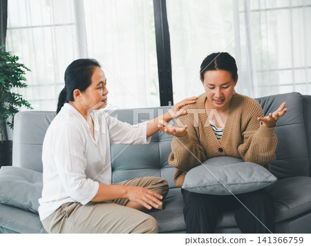 Asian mother listening and comforting adult daughter during serious conversation on sofa at home, concept of family counseling, emotional support, trust, and mental wellness. Asian mother listening and comforting adult daughter during serious conversation on sofa at home, concept of family counseling, emotional support, trust, and mental wellness. 141366759
