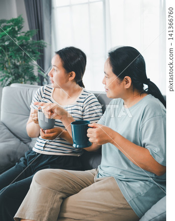 Asian mother and adult daughter sitting on sofa with cups at home, having conversation and pointing outside, concept of family communication, support, care, and trust. Asian mother and adult daughter sitting on sofa with cups at home, having conversation and pointing outside, concept of family communication, support, care, and trust. 141366760
