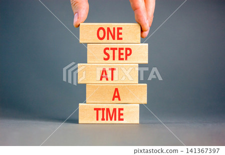 One step at a time symbol. Concept words One step at a time on beautiful wooden blocks. Beautiful grey background. Businessman hand. Business and one step at a time concept. Copy space. One step at a time symbol. Concept words One step at a time on beautiful wooden blocks. Beautiful grey background. Businessman hand. Business and one step at a time concept. Copy space. 141367397
