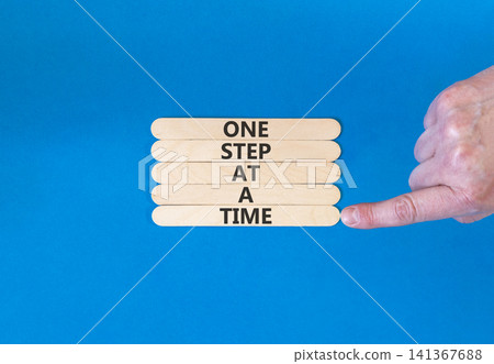 One step at a time symbol. Concept words One step at a time on beautiful wooden sticks. Beautiful blue background. Businessman hand. Business and one step at a time concept. Copy space. One step at a time symbol. Concept words One step at a time on beautiful wooden sticks. Beautiful blue background. Businessman hand. Business and one step at a time concept. Copy space. 141367688