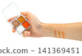 Hand holding a small four-color eyeshadow palette with orange, brown, and glitter shades. Makeup swatches applied to the forearm. Professional beauty product photography isolated. 141369451