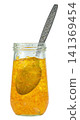 Transparent glass jar filled with orange marmalade preserve and a decorative silver spoon inside Isolated. Sweet citrus fruit jam for breakfast. Professional food photography. 141369454