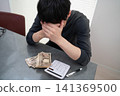 一名男子雙手抱頭，前方是一堆錢和一台計算機。 141369500