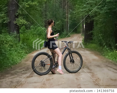 Caucasian woman riding a mountain bike on a dirt trail in a green forest. Active leisure lifestyle, outdoor exercise, and summer countryside travel. Sporty girl cycling in nature. Caucasian woman riding a mountain bike on a dirt trail in a green forest. Active leisure lifestyle, outdoor exercise, and summer countryside travel. Sporty girl cycling in nature. 141369758