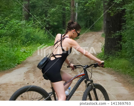 Caucasian woman riding a mountain bike on a dirt trail in a green forest. Active leisure lifestyle, outdoor exercise, and summer countryside travel. Sporty girl cycling in nature. Caucasian woman riding a mountain bike on a dirt trail in a green forest. Active leisure lifestyle, outdoor exercise, and summer countryside travel. Sporty girl cycling in nature. 141369763