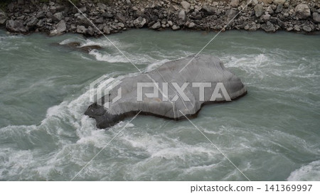 Large gray boulder partially submerged in rushing turquoise-white water and foam, geology concept. Large gray boulder partially submerged in rushing turquoise-white water and foam, geology concept. 141369997
