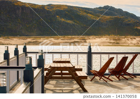 terrace of a house by the water on a sunny autumn day in Teriberka, sandy beach, mountain view, tables and chairs on the terrace, deck chairs, Murmansk region, Russia, Kola terrace of a house by the water on a sunny autumn day in Teriberka, sandy beach, mountain view, tables and chairs on the terrace, deck chairs, Murmansk region, Russia, Kola 141370058