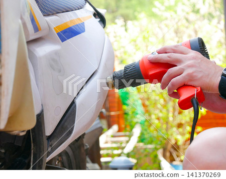 Man using heat gun to repair plastic motorcycle bodywork. Mechanic warming up and reshaping scratched fairing for vehicle restoration. Automotive maintenance and DIY garage repair service. Man using heat gun to repair plastic motorcycle bodywork. Mechanic warming up and reshaping scratched fairing for vehicle restoration. Automotive maintenance and DIY garage repair service. 141370269