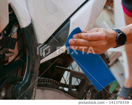 Man repairing motorcycle using sandpaper. Mechanic sanding plastic motorcycle frame to polish surface. Vehicle maintenance and restoration service. DIY hobby work on dirt bike in garage. Man repairing motorcycle using sandpaper. Mechanic sanding plastic motorcycle frame to polish surface. Vehicle maintenance and restoration service. DIY hobby work on dirt bike in garage. 141370339