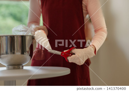 Baker in burgundy apron cutting plastic piping bag with red scissors while preparing cake batter in stainless bowl Baker in burgundy apron cutting plastic piping bag with red scissors while preparing cake batter in stainless bowl 141370400