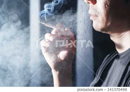 一名男子手裡拿著香煙,嘴裡被白煙籠罩。 一名男子手裡拿著香煙,嘴裡被白煙籠罩。 141370499