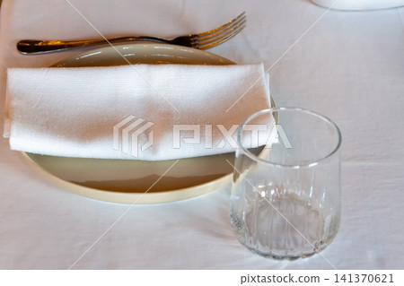 Restaurant table setting with clear drinking glass white cloth napkin and ceramic plate on a white tablecloth Restaurant table setting with clear drinking glass white cloth napkin and ceramic plate on a white tablecloth 141370621