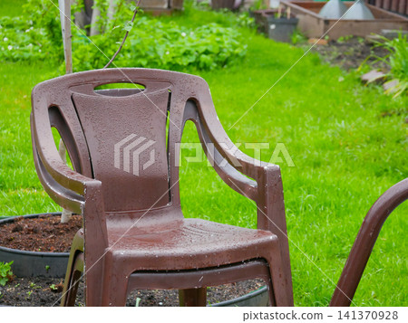 Brown plastic chair standing on green lawn in garden after rain. Wet patio furniture covered with water drops. Backyard leisure and outdoor relaxation concept in spring season. Brown plastic chair standing on green lawn in garden after rain. Wet patio furniture covered with water drops. Backyard leisure and outdoor relaxation concept in spring season. 141370928