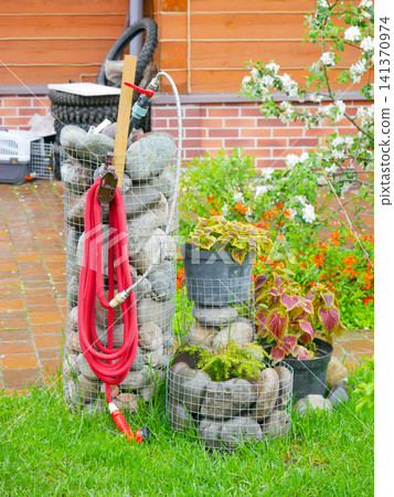 Gabion structures made of wire mesh and stones used for garden decoration and hose storage. Functional landscaping element with red water hose in backyard. Creative outdoor design idea. Gabion structures made of wire mesh and stones used for garden decoration and hose storage. Functional landscaping element with red water hose in backyard. Creative outdoor design idea. 141370974