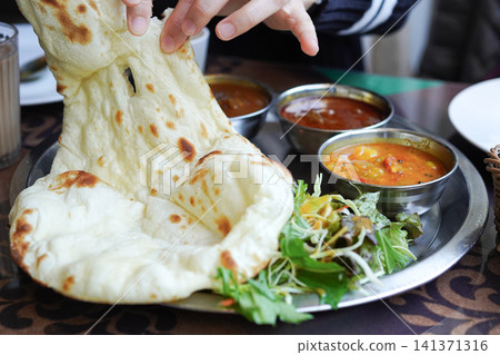 The hands of a person tearing off pieces of freshly baked naan and eating curry at an Indian restaurant. The hands of a person tearing off pieces of freshly baked naan and eating curry at an Indian restaurant. 141371316