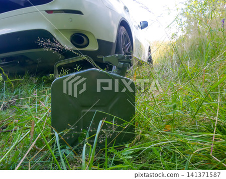 Green metal fuel canister standing in tall grass next to white modern car. Emergency gasoline supply for stranded vehicle in countryside. Travel problem and road safety concept. 141371587