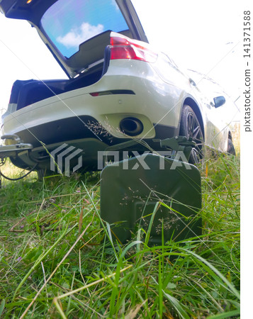 Green metal jerrycan standing on grass field near white car with open trunk. Emergency fuel supply for vehicle trip. Roadside assistance and automotive travel safety concept. Green metal jerrycan standing on grass field near white car with open trunk. Emergency fuel supply for vehicle trip. Roadside assistance and automotive travel safety concept. 141371588