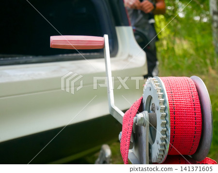 Man using manual boat trailer winch to secure watercraft. Hand operated strap mechanism for loading boat onto vehicle. Transportation hardware and marine equipment safety gear concept. Man using manual boat trailer winch to secure watercraft. Hand operated strap mechanism for loading boat onto vehicle. Transportation hardware and marine equipment safety gear concept. 141371605