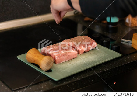 Hand Sprinkling Pepper on Seasoned Raw Pork Steaks Board 141371616