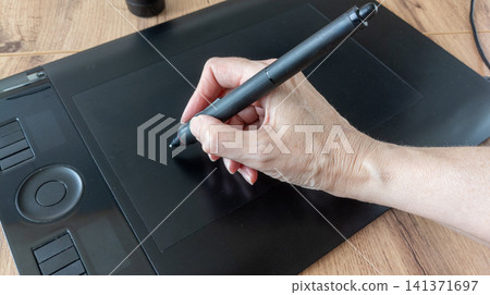 Artist hand wearing a black two-finger artist glove draws with a digital stylus pen on a black graphic tablet. Creative digital illustration process Artist hand wearing a black two-finger artist glove draws with a digital stylus pen on a black graphic tablet. Creative digital illustration process 141371697