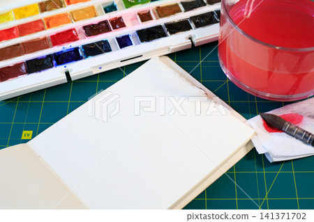 Open sketchbook, watercolor paints, brush, and water on a green mat. Art tools for watercolor painting 141371702