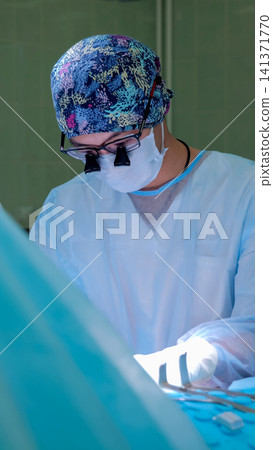 Surgeon in patterned scrub cap, mask, magnifying loupes, and sterile gown performing delicate operation under bright surgical lights inside operating room Surgeon in patterned scrub cap, mask, magnifying loupes, and sterile gown performing delicate operation under bright surgical lights inside operating room 141371770
