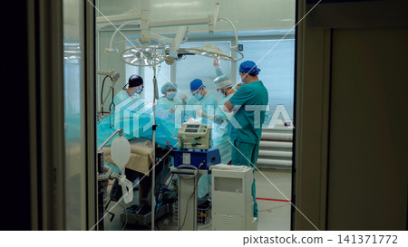 Surgical team working in sterile operating room with blue drapes, overhead lights, medical monitors, equipment, and glass doorway view during hospital procedure Surgical team working in sterile operating room with blue drapes, overhead lights, medical monitors, equipment, and glass doorway view during hospital procedure 141371772