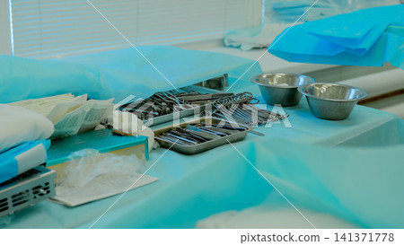 Sterile surgical instruments arranged on blue operating room drape with stainless steel bowls, metal trays, forceps, scissors, clamps, folded towels, and gauze prepared for clinical procedure Sterile surgical instruments arranged on blue operating room drape with stainless steel bowls, metal trays, forceps, scissors, clamps, folded towels, and gauze prepared for clinical procedure 141371778