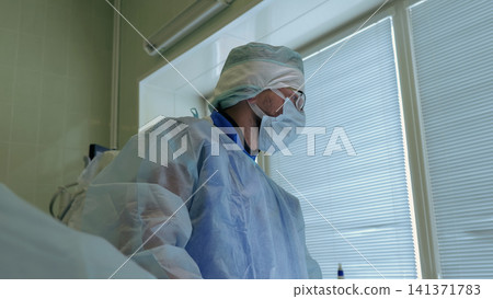 Medical professional wearing protective gown, face mask, surgical cap, and glasses standing beside hospital bed near window blinds in clean clinical room with soft daylight Medical professional wearing protective gown, face mask, surgical cap, and glasses standing beside hospital bed near window blinds in clean clinical room with soft daylight 141371783