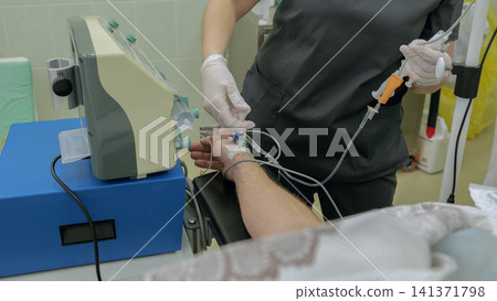 Gloved nurse preparing intravenous infusion on patient forearm in clinic treatment room with catheter tubing, sterile dressing, drip stand, pump, monitor, and reclining chair nearby Gloved nurse preparing intravenous infusion on patient forearm in clinic treatment room with catheter tubing, sterile dressing, drip stand, pump, monitor, and reclining chair nearby 141371798