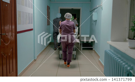 Medical staff transporting patient on wheeled hospital bed through bright corridor with pale teal walls, tiled floor, radiator, windows, handrails, and clinical interior lighting Medical staff transporting patient on wheeled hospital bed through bright corridor with pale teal walls, tiled floor, radiator, windows, handrails, and clinical interior lighting 141371799