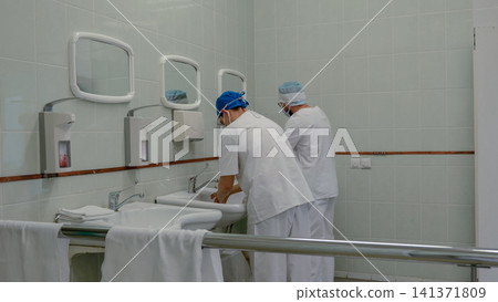 Medical staff washing hands in tiled hospital scrub room before surgery, wearing masks, caps, and white uniforms beside stainless steel sinks, mirrors, soap dispensers, and bright clinical lighting Medical staff washing hands in tiled hospital scrub room before surgery, wearing masks, caps, and white uniforms beside stainless steel sinks, mirrors, soap dispensers, and bright clinical lighting 141371809