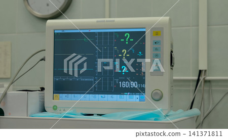 Bedside patient monitor showing flat cardiac waveform and numeric vital readings on wheeled trolley in quiet clinical room with blue surgical drape, tiled wall, soft fluorescent light, and medical 141371811