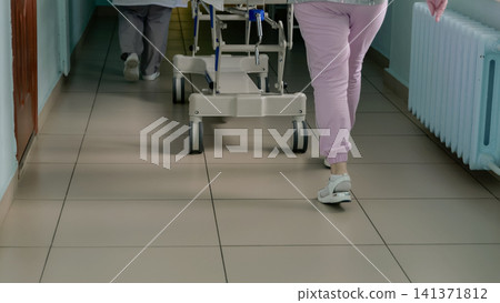 Medical stretcher wheels and staff lower legs moving through hospital corridor with tiled floor, pale green walls, radiators, clinical lighting, and urgent patient transport atmosphere 141371812