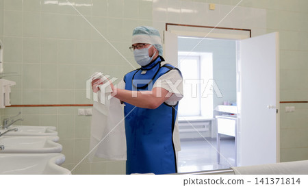 Masked medical worker wearing protective blue lead apron and cap stands in clean tiled hospital washroom while holding white coat near sinks with open doorway and soft clinical light Masked medical worker wearing protective blue lead apron and cap stands in clean tiled hospital washroom while holding white coat near sinks with open doorway and soft clinical light 141371814