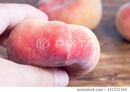 Close up shot of fingers holding a ripe donut peach with other peaches out of focus. 141372164