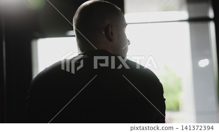 Backlit rear three quarter view of man standing indoors by bright window with shoulders squared and head turning slightly, creating moody contrast, visible skin texture, and calm reflective mood Backlit rear three quarter view of man standing indoors by bright window with shoulders squared and head turning slightly, creating moody contrast, visible skin texture, and calm reflective mood 141372394