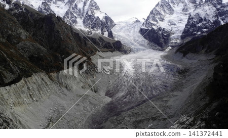 Elevated panorama of alpine glacier tongue descending between jagged snow covered peaks, with exposed moraine forefield, compacted blue ice, dark debris streaks, rugged rock walls and overcast Elevated panorama of alpine glacier tongue descending between jagged snow covered peaks, with exposed moraine forefield, compacted blue ice, dark debris streaks, rugged rock walls and overcast 141372441