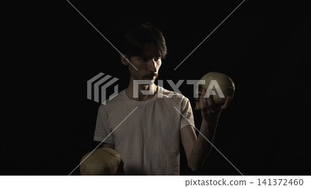 Serious young man holding two human skulls in dramatic studio lighting Serious young man holding two human skulls in dramatic studio lighting 141372460