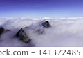 Jagged mountain peaks rising through drifting cloud deck beneath clear blue sky, dramatic alpine panorama with dark rock summits, mist, vast horizon, serene highland scale 141372485
