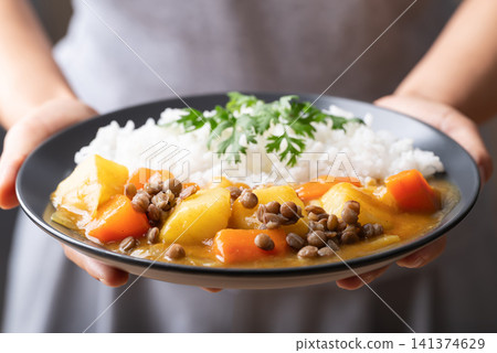 Japanese curry with lentil, carrot, potato, onion and cooked rice on plate holding by woman hand Japanese curry with lentil, carrot, potato, onion and cooked rice on plate holding by woman hand 141374629