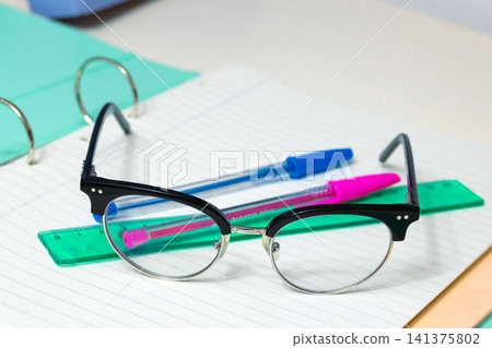School supplies including glasses, two colorful pens, and a ruler laying on a lined open notebook, symbolizing readiness for learning, studying, and academic endeavors School supplies including glasses, two colorful pens, and a ruler laying on a lined open notebook, symbolizing readiness for learning, studying, and academic endeavors 141375802
