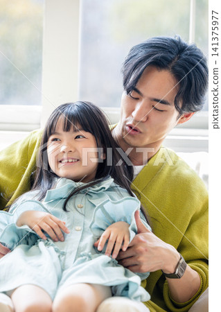 A little girl is sitting next to her father in the living room, smiling gently. 141375977