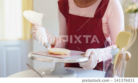 Young Asian woman decorating cake with icing on stand in bright home kitchen while baking 141376831