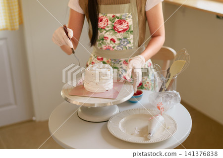 Asian woman pastry chef spreading white frosting on layered cake using offset spatula in bright home kitchen Asian woman pastry chef spreading white frosting on layered cake using offset spatula in bright home kitchen 141376838
